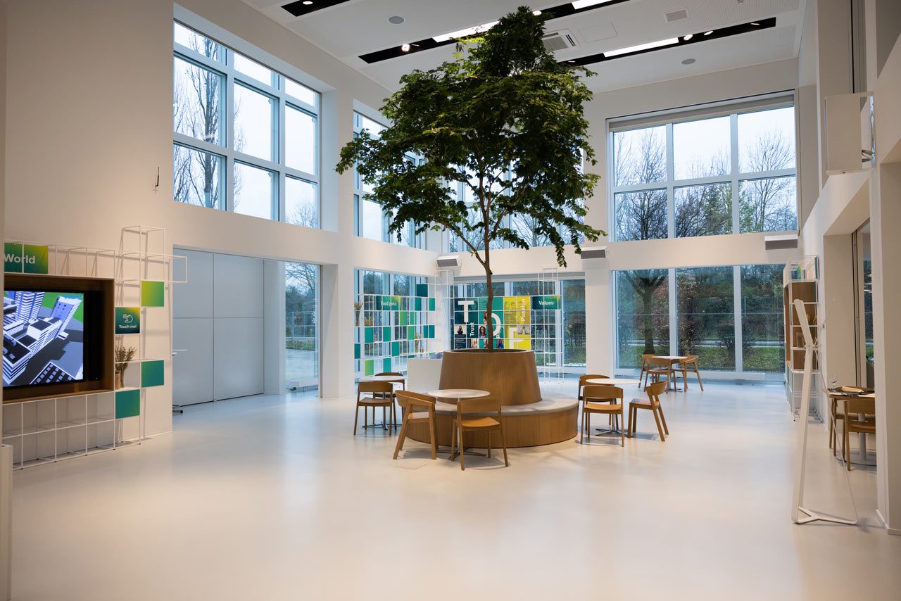 Hoher heller Raum mit großem Baum in der Mitte, umgeben von Sitzgruppen und raumhohen Fenstern