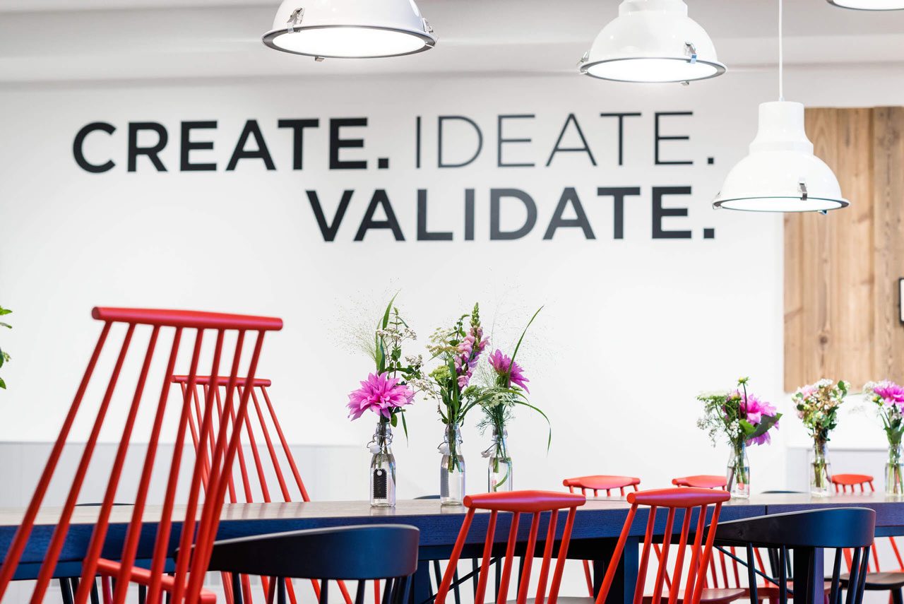 Workshopraum mit roten Stühlen an dunklen Tischen, an der Wand der Schriftzug „Create. Ideate. Validate.“