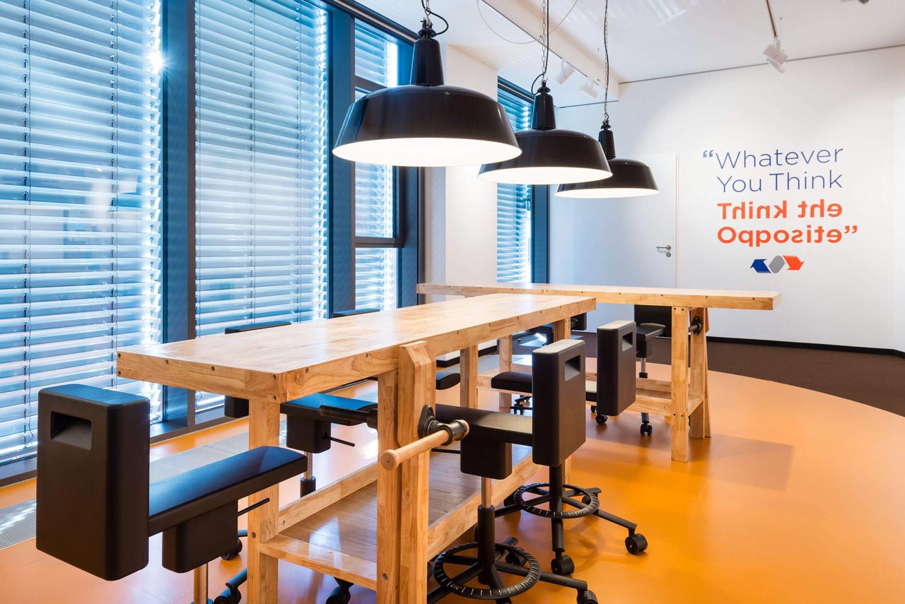 Werkbank aus Holz mit Hockern und Bürostühlen, an der Wand der Schriftzug „Whatever you think, think the opposite“