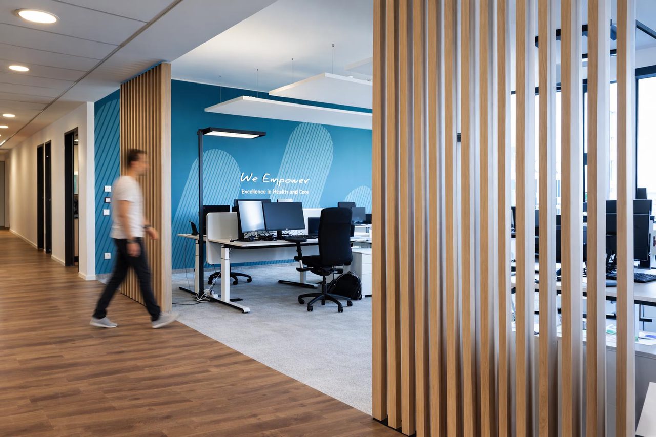 Blick durch einen Gang: rechts eine Wand aus Holzlamellen, links Arbeitsplätze vor blauer Wand