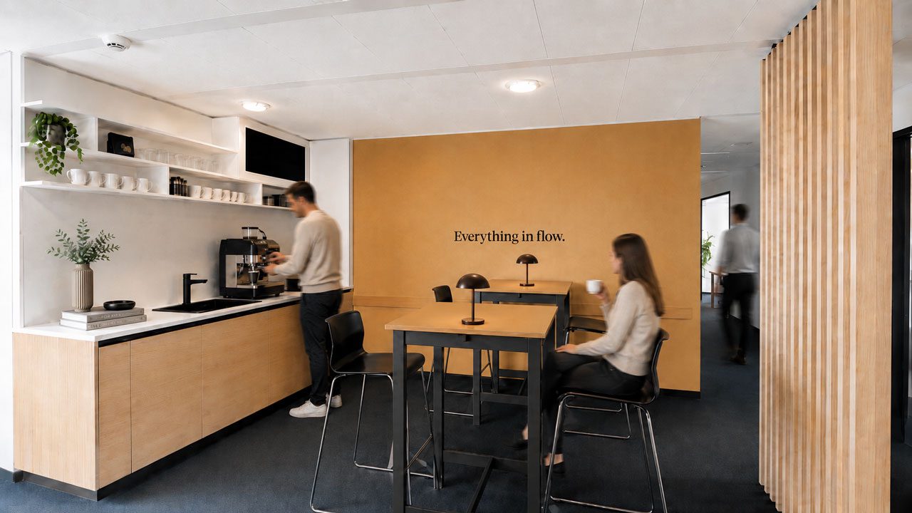 Küchenzeile mit Siebträgermaschine, daneben ein Stehtisch mit zwei Personen vor einer ockerfarbenen Wand mit dem Schriftzug „Everything in flow“