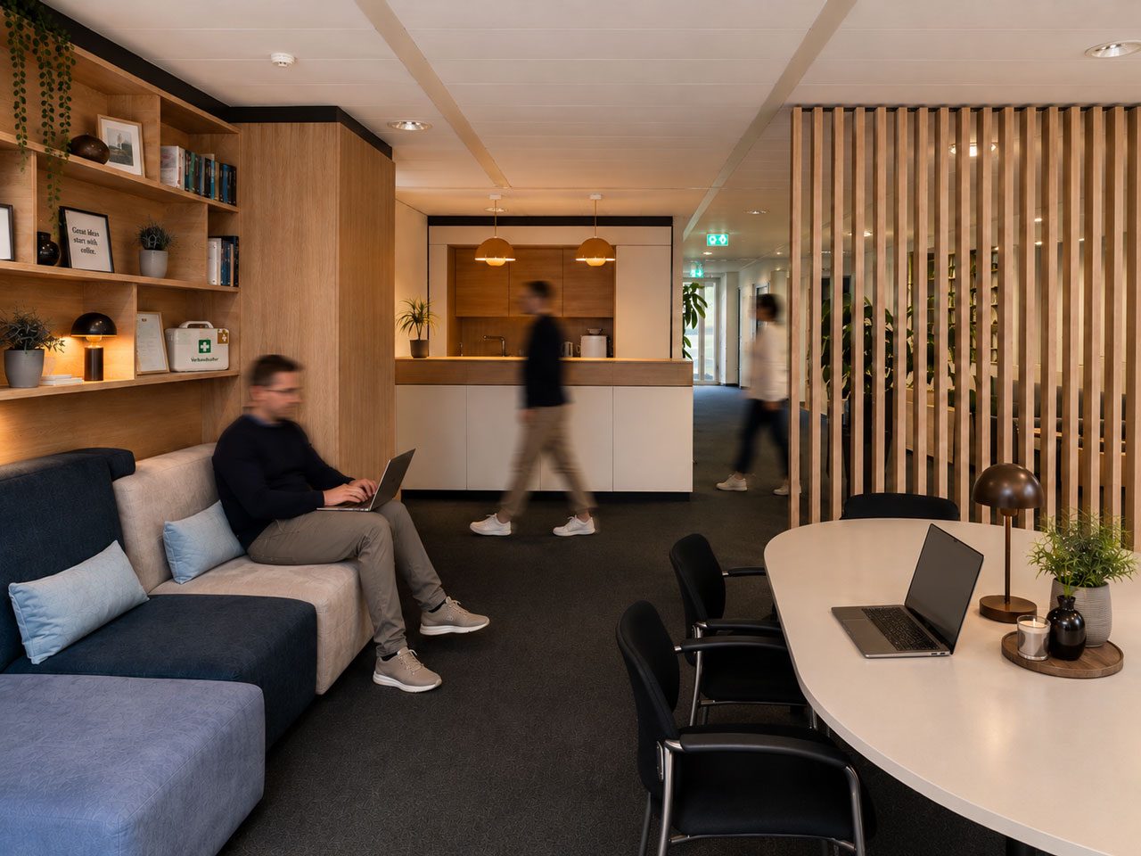 Lounge mit grauem Sofa, Regalwand und ovalem Besprechungstisch, im Hintergrund der Empfang hinter Holzlamellen