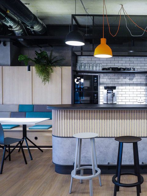 Café-Bar aus hellem Holz mit Barhockern, darüber gelbe und weiße Pendelleuchten und hängende Pflanzen