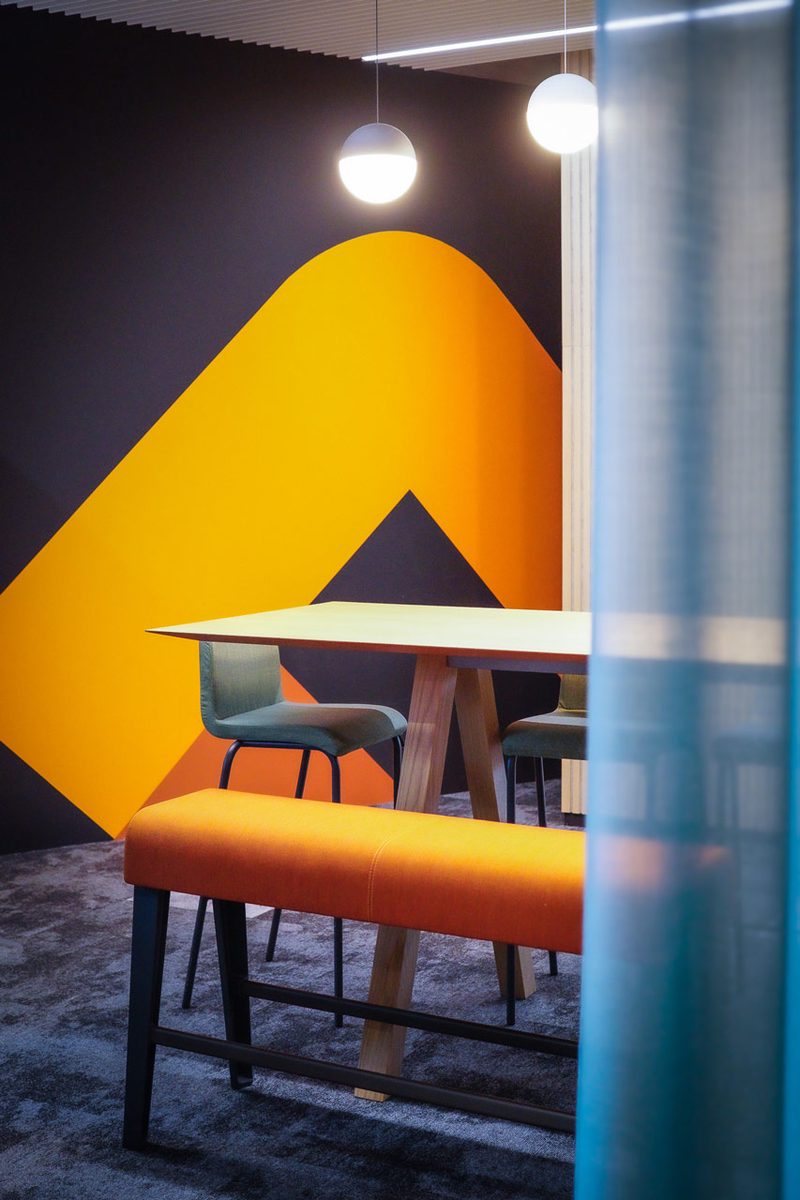 Sitznische mit oranger Polsterbank und Holztisch vor einer Wandgrafik in Orange und Gelb