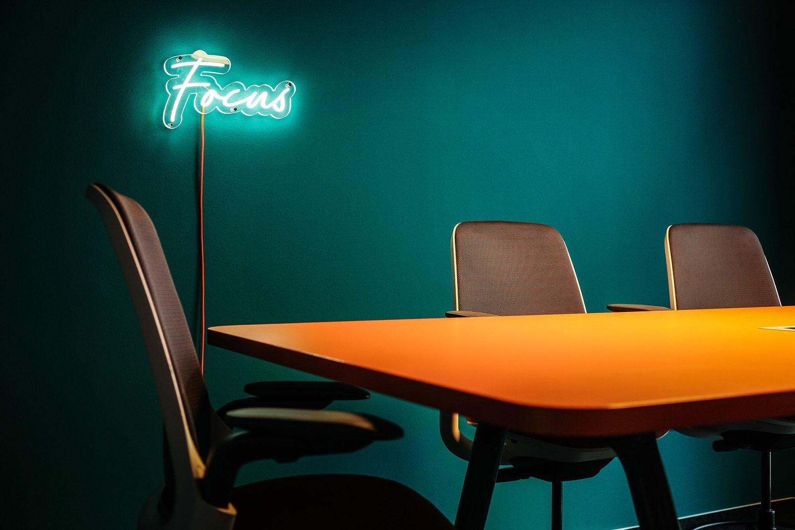 Fokusraum mit orangem Tisch, Holzstühlen und der Leuchtschrift „Focus“ an der petrolfarbenen Wand