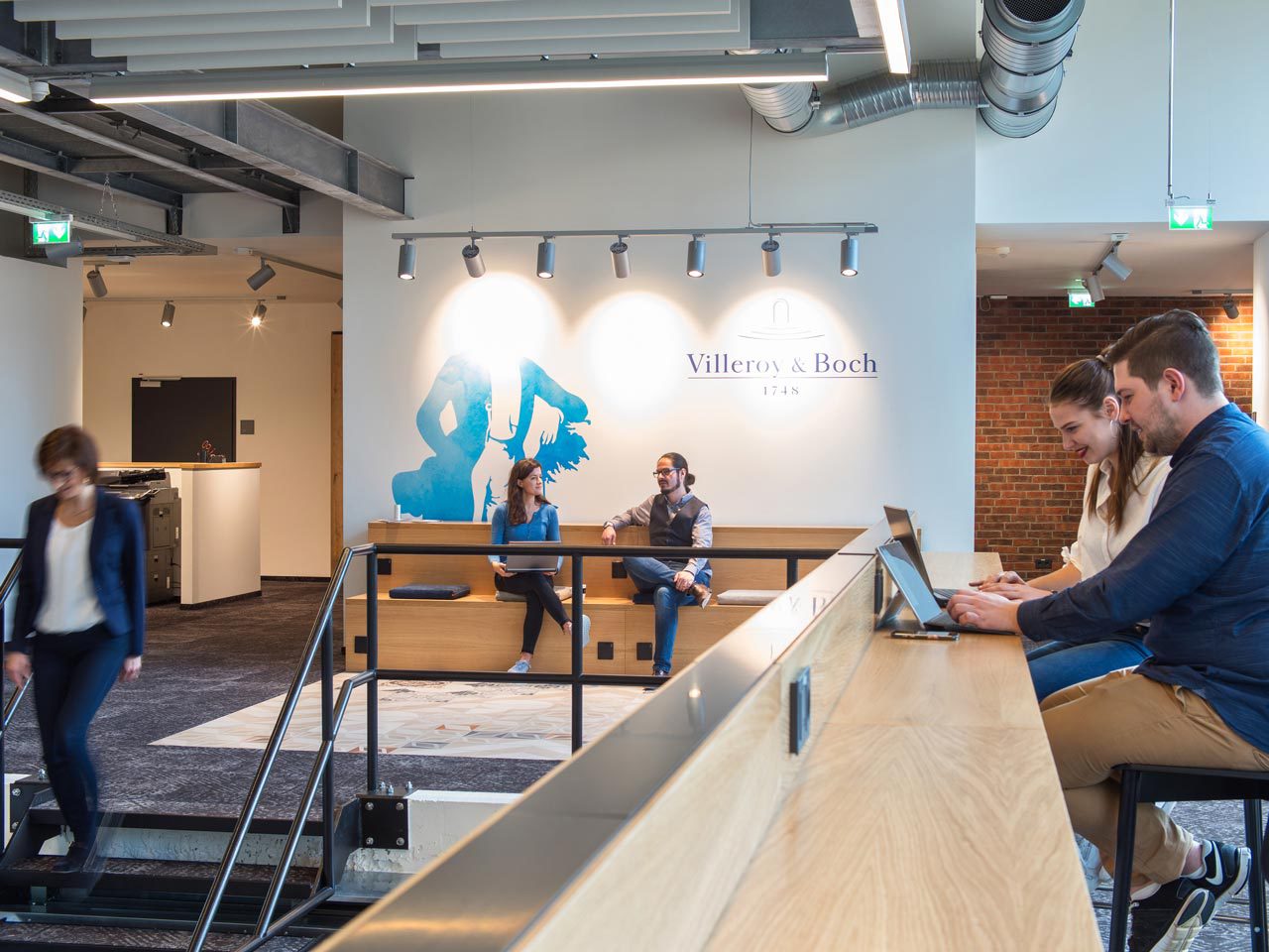 Caféartiger Arbeitsbereich mit Tresen aus hellem Holz, Personen an Laptops, an der Wand eine blaue Grafik mit dem Villeroy-&-Boch-Schriftzug