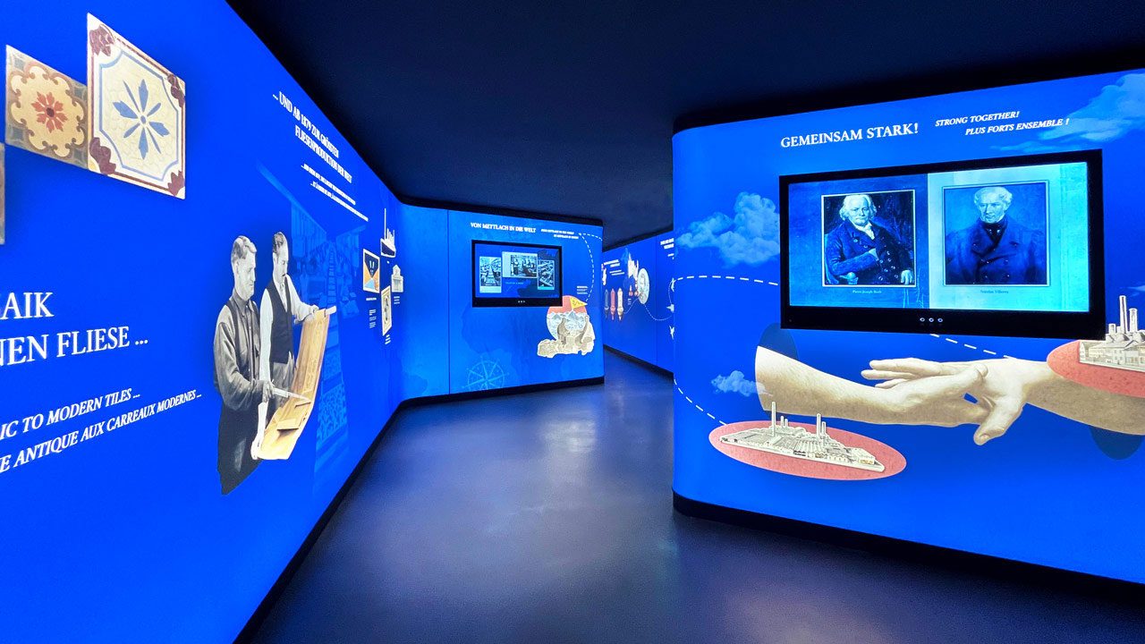 Blau ausgeleuchteter Raum mit Bildschirmen, Ausstellungstischen und Figuren an der Wand